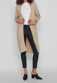 Cárdigan de punto beige con mangas largas y bolsillos laterales, combinado con pantalones de cuero negros y mules negros de tacón.