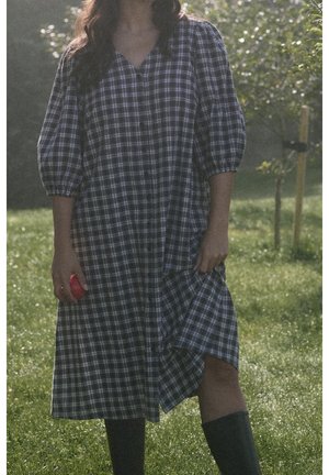 Femme portant une longue robe à carreaux et des bottes noires, debout sur l'herbe, tenant un petit fruit rouge d'une main, en plein air avec des arbres.