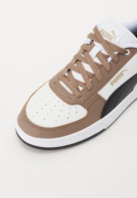 Braun-weiße Sneaker mit einem strukturierten Leder-Obermaterial, perforiertem Zehenbereich und beigen Schnürsenkeln. Verfügt über einen schwarzen Akzent und goldene Markenkennzeichnung.