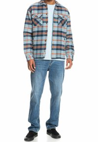 Man wearing blue, orange, and navy plaid button-up shirt, light blue t-shirt, blue jeans, and black slip-on shoes, standing against white background.