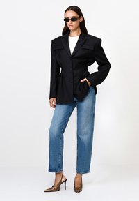 Naine, kes kannab ülemõõdulist musta bleiserit, valget pusa, siniseid teksaseid, leopardimustriga kõrge kontsaga kingi ja musti päikeseprille, seisab valge tausta vastu.