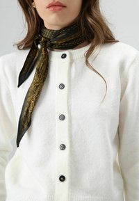 Cardigan blanc en maille avec un col côtelé, boutons noirs, accompagné d'une écharpe noire et dorée à motif, nouée autour du cou.