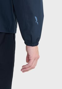 Laivaston vedenpitävä takki, jossa on joustavat hihansuut. Vaaleansininen logo korostaa hihaa. Kangas näyttää sileältä ja kestävältä.