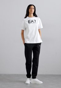 Hvit kortermet T-skjorte med svart brodert EA7-logo, kombinert med svarte bukser og hvite sneakers. Enkel, uformell design.