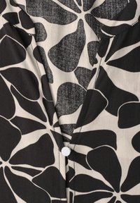 Blouse noire et beige avec un grand motif floral abstrait et un petit bouton blanc rond au col.