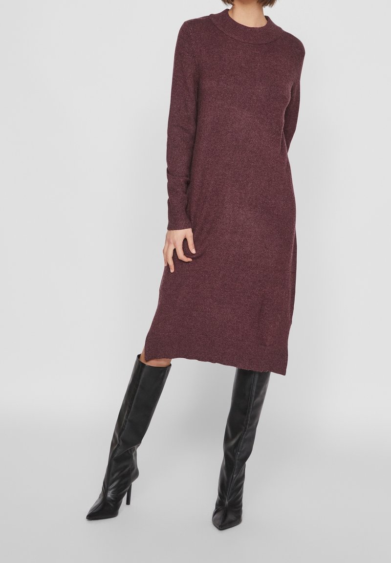Femme portant une robe en maille bordeaux longueur genoux à manches longues, associée à des bottes noires à talons hauts montant jusqu'aux genoux, debout devant un fond uni.