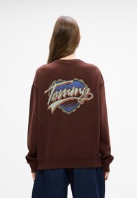 Sudadera marrón con un gran logo colorido de "Tommy" que incluye un diseño de corazón y detalles de cinta, fabricada con una tela suave y texturizada.