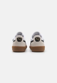 Sneaker mit einem weißen Obermaterial, grauem Wildleder-Fersenbereich und einer braunen Gummisohle. Verfügt über ein goldenes Logo auf der Rückseite und schwarze seitliche Akzente.