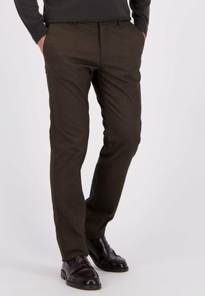 Man in een donkerbruin slim-fit broek en zwarte leren instappers, met de handen in zijn zakken, staande tegen een effen lichte achtergrond.