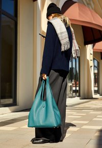 Turkusowa skórzana torba typu tote o luźnym kształcie, gładkiej teksturze i dwóch wydłużonych uchwytach; posiada złote logo i stoi na czterech ochronnych nóżkach.