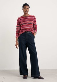 Pulover rayat roșu și roz, purtat cu pantaloni large din denim închis. Puloverul are mâneci lungi și o croială lejeră; pantalonii au talie înaltă și cusături vizibile.