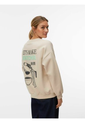 Donna che indossa una felpa beige con testo "Let's make matcha" e grafiche per la preparazione del tè matcha sulla schiena, che guarda oltre la spalla.