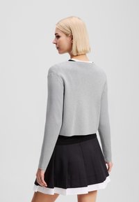 Maglione grigio corto con maniche lunghe, texture in maglia, abbinato a una gonna nera plissè con uno strato sottostante bianco.
