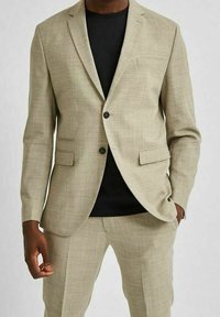 Ljusbeige kostym med texturerat tyg, enknappsstängning, slagkragar och två framfickor, bärs över en svart skjorta.