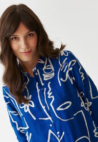 Camicia blu realizzata in tessuto con una superficie liscia, caratterizzata da disegni astratti in bianco lungo tutta la superficie, con chiusura a bottoni e colletto.