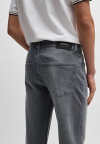 Grå denim bukser med slim fit, der har baglommer og en tekstureret læderlogo patch mærket "BOSS." Glat finish med et minimalistisk design.