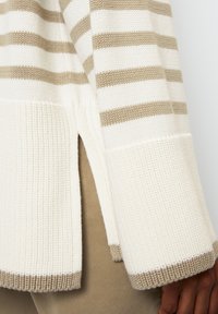 Gestrickter Pullover mit weißen und hellbeigen Streifen, ausgestattet mit Seitenschlitzen, geripptem Saum und strukturierter Stoffqualität in einer lockeren Passform.