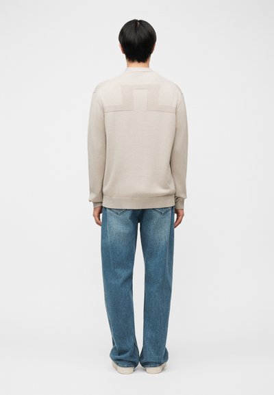 Άτομο με μαύρα μαλλιά που φοράει μπεζ πουλόβερ με υφή γεωμετρικού σχεδίου στην πλάτη, μπλε τζιν και ανοιχτόχρωμα παπούτσια, στέκεται γυρισμένο προς τα πίσω.