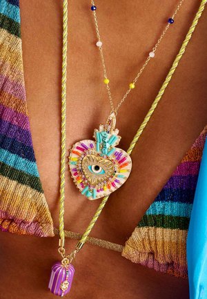 Goldfarbener herzförmiger Anhänger mit bunten Verzierungen, türkisfarbenen Details und einem Augenmotiv. Enthält einen lavendelfarbigen dekorativen Charm.