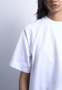 Άτομο που φοράει απλό λευκό κοντομάνικο μπλουζάκι, ορατό από τους ώμους έως το μέσο του κορμού, με ουδέτερο ανοιχτό γκρι φόντο.