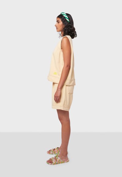 Top senza maniche beige e pantaloni corti abbinati, con una tasca e ricami gialli. Sandali con motivo floreale colorato completano il look.