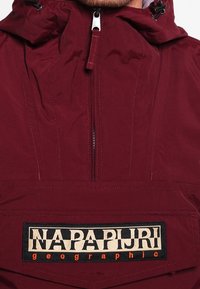 Bordeaux capuchonjack met een rits aan de voorkant, een grote zak met een 'NAPAPIJRI' logo patch en een gestructureerde stof.