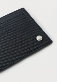 Μαύρο πορτοφόλι από δερματίνη με υφή, με τρεις θήκες για κάρτες και ένα μικρό ασημί στρογγυλό λογότυπο σε λευκό φόντο.