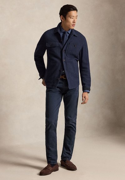 Vyras vilki tamsiai mėlyną švarką ant languotos marškinių ir kaklaraiščio, tamsias kelnes ir rudas laisvalaikio batus, stovi ir žiūri į kairę.