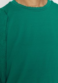 Pull vert avec un col à festons et des détails en dentelle texturée sur les manches. Le tissu semble lisse et extensible.