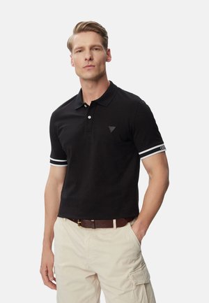 Hombre con camiseta tipo polo negra de manga corta con rayas blancas en las mangas y pantalones cargo beige, de pie con una mano en el bolsillo.