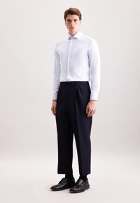 Camicia bianca con colletto, abbinata a pantaloni pleated blu scuro e scarpe nere stringate. Tessuto liscio con design aderente.