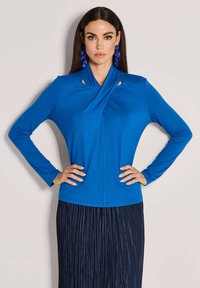 Blaues Langarmoberteil mit einem einzigartigen Drapierungs-Ausschnitt und Cut-out-Details. Weicher Stoff, figurbetontes Design, kombiniert mit einem plissierten marineblauen Rock.