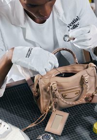 Person trägt weiße Handschuhe und einen weißen Laborkittel und untersucht mit einer Lupe das Innere einer beigen Lederhandtasche.
