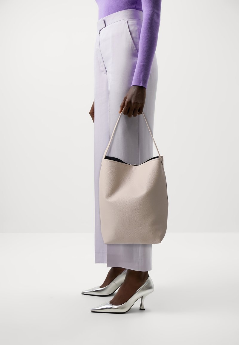 Personne portant un haut à manches longues lavande, un pantalon violet clair, des talons hauts argentés, tenant un sac à main beige texturé par sa sangle.