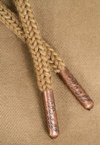 Geflochtene, hellbraune Kordelenden, abgeschlossen mit braunen Metallspitzen, die mit dekorativem Text auf passendem Stoffhintergrund geprägt sind.