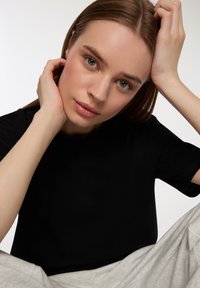 Czarny bawełniany t-shirt z krótkim rękawem, okrągłym dekoltem i luźnym krojem. Modelka siedzi, ubrana w jasnoszare spodnie, z prostymi brązowymi włosami.