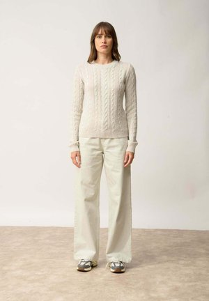 Suéter de punto trenzado en crema claro con cuello redondo, combinado con pantalones de pierna ancha en beige. El modelo lleva zapatillas en gris y blanco. Fondo sencillo.