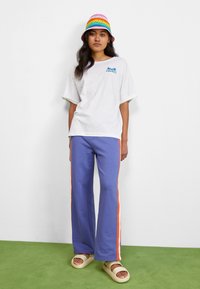 Jonge vrouw in een witte T-shirt, paarse broek met oranje en witte zijstrepen, crèmekleurige sandalen en een regenboogkleurige gehaakte bucket hat.