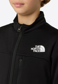 Black fleece jacket with a high collar, featuring a front zip and a white logo. Textured fabric with a zippered pocket detail.