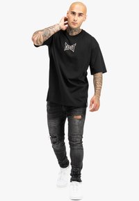Juodas trumpomis rankovėmis T-shirt su baltu "TAPOUT" logotipu ant krūtinės, kartu su dėvėtais juodais džinsais ir baltais sportbačiais.