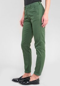 Chinos en coton vert avec une coupe slim, dotés d'une fermeture à bouton, de quatre poches et d'un ourlet roulé. Associés à des mocassins noirs en vernis.
