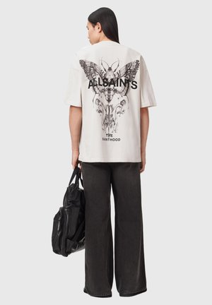 Henkilö, jolla on päällään vaaleanbeige ylisuurikokoinen t-paita, jossa on musta perhonen ja kallo -kuvio sekä teksti "ALLSAINTS LONDON" takana, ja joka pitää mustaa laukkua.