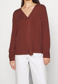 Top marron à manches longues avec un décolleté en V, en tissu doux. Présente des manches froncées et une coupe ample. Assorti avec un pantalon blanc.