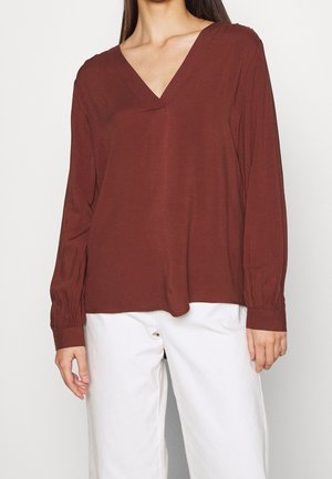Top marron à manches longues avec un décolleté en V, en tissu doux. Présente des manches froncées et une coupe ample. Assorti avec un pantalon blanc.