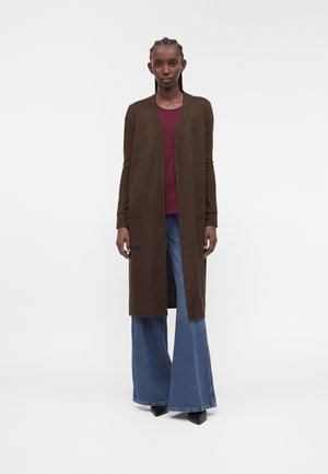 Femme grande aux cheveux tressés portant un long cardigan marron, un haut bordeaux, un jean bleu à larges jambes et des chaussures noires pointues, debout sur fond blanc.