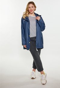 Mörkblå vattentät jacka med huva, guldfärgade knappar och fickor med dragkedja, bärs över en randig skjorta och grå jeans med vita sneakers.