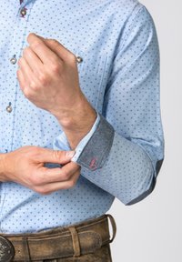 Muž v košili s vzorem v světle modré barvě zapíná manžetu, nosí hnědý kožený opasek s ozdobnou sponou, na neutrálním pozadí.