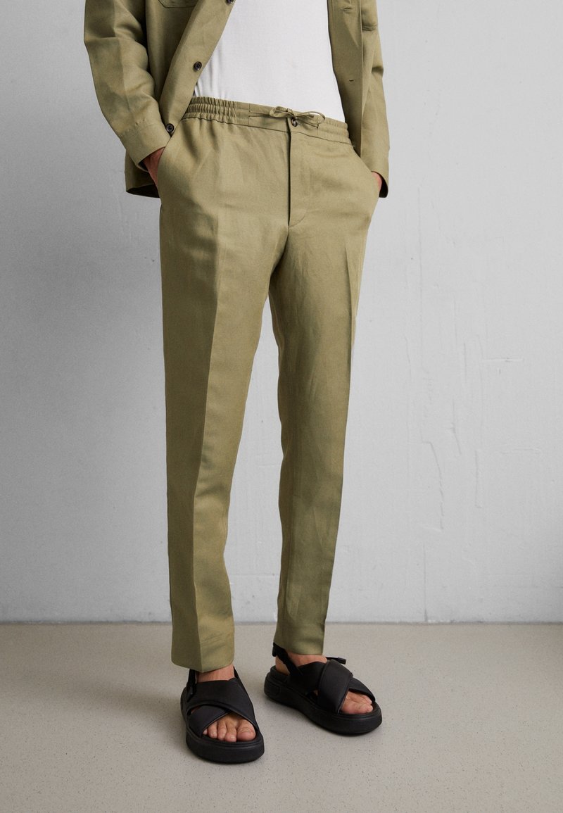 Homme portant un pantalon beige sur mesure avec taille élastique et des sandales noires ouvertes aux orteils, debout contre un mur gris clair uni.