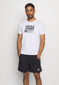 White cotton t-shirt featuring "JACK & JONES PERFORMANCE" printed in bold black text, paired with black athletic shorts.