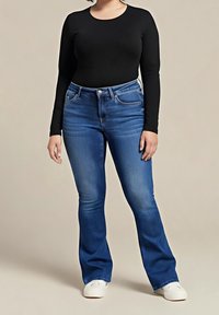 Vaqueros acampanados de color azul con un lavado medio, que cuentan con un diseño de cinco bolsillos y cierre de botones, combinados con una camiseta negra de manga larga y zapatos blancos.